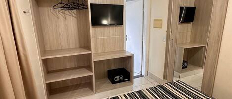 A hotel room with a bed, a TV mounted on the wall, a wardrobe with shelves, and a small cabinet.
