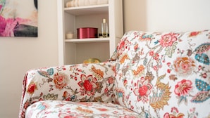 Un sofá con estampado floral, un estante blanco con una maceta roja y una botella de vidrio, y un cuadro en la pared.