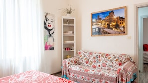 Una habitación con un sofá estampado con motivos florales, una cama con colcha estampada, una estantería y un cuadro enmarcado de un paisaje urbano.
