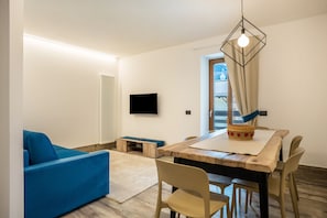 Een moderne woonkamer met een blauwe bank, een houten eettafel met stoelen, een aan de muur bevestigde televisie en een raam met gordijnen.