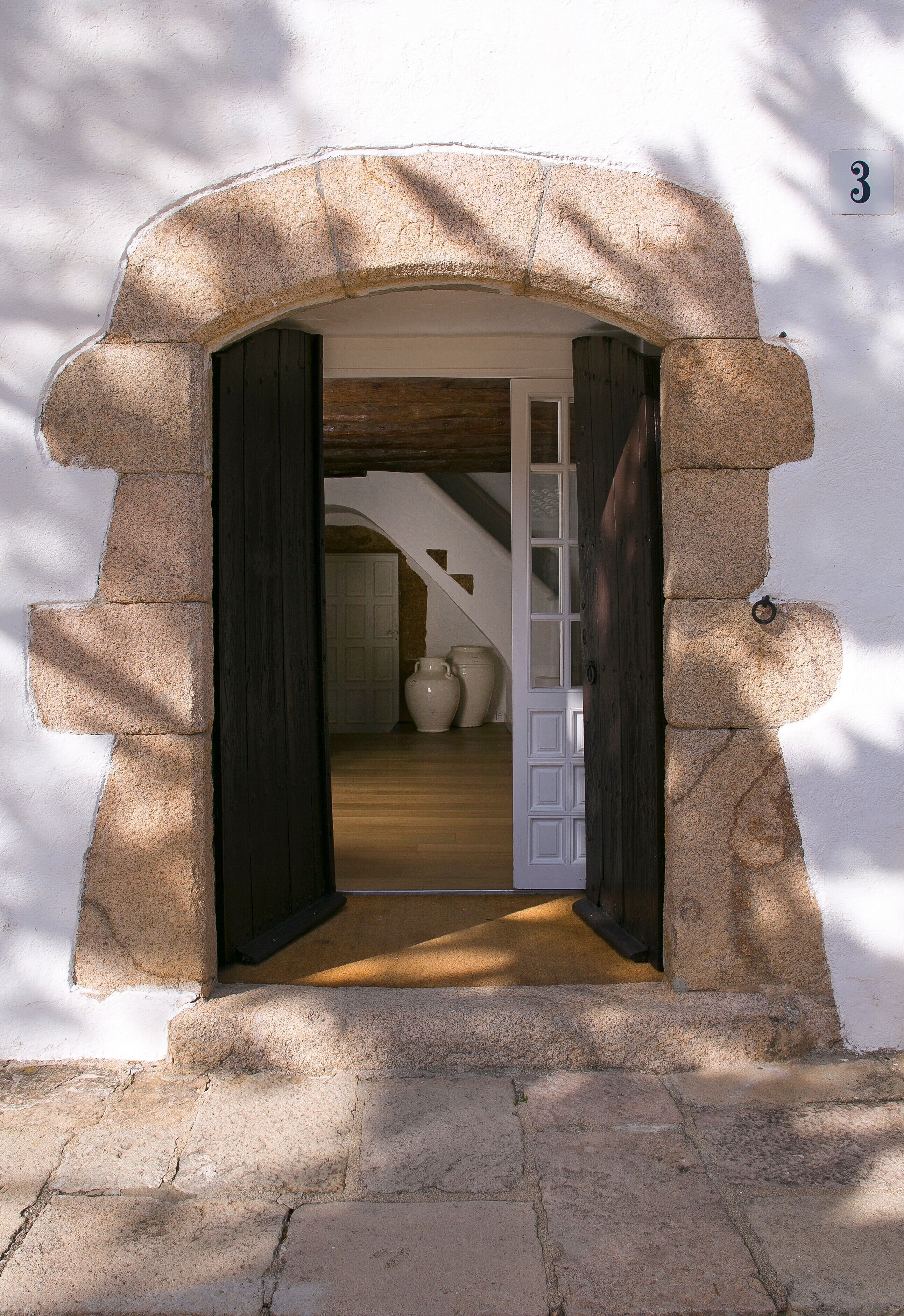 Un arco de piedra da acceso a una habitación con suelo de madera, una puerta blanca y un jarrón grande.