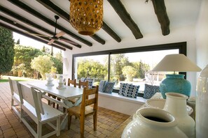 Un área de comedor al aire libre con una mesa blanca, sillas de madera, un ventilador de techo y vistas a la vegetación a través de grandes ventanales.