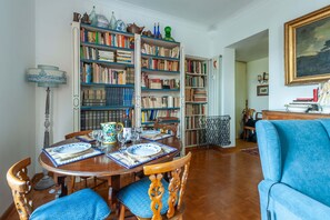 A dining area with a round table set for four, surrounded by chairs with blue cushions. A large bookshelf filled with books and decorative items is visible in the background.