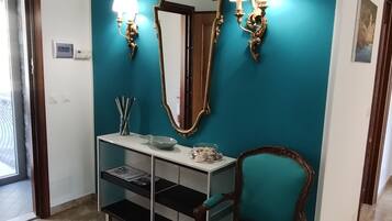 Une pièce aux murs de couleur turquoise, avec une chaise en bois, une console blanche ornée d’un miroir et de deux appliques murales, et une porte donnant sur l’extérieur.