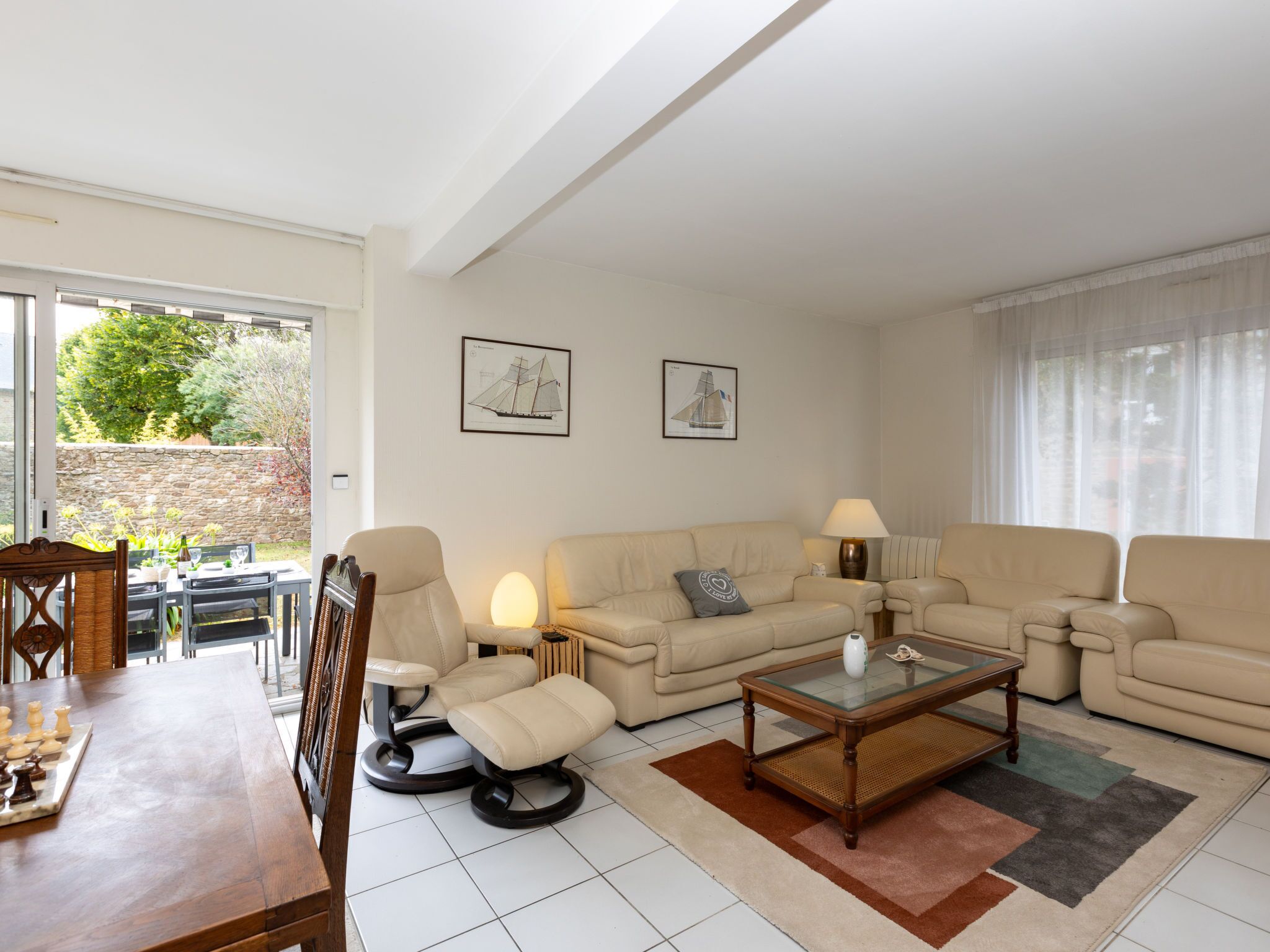Ein Wohnzimmer mit einer Couch, Sesseln, einem Couchtisch und einem Schachbrett. An den Wänden hängen zwei gerahmte Bilder, ein Fenster mit Vorhängen und ein Blick auf einen Garten.
