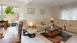 Een woonkamer met een bank, fauteuils, een bijzettafeltje en een schaakbord. Twee ingelijste foto's hangen aan de muur, een raam met gordijnen en uitzicht op een tuin.