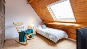 Een knus zolderkamer met schuine wanden, een enkel bed, een schommelstoel, een klein nachtkastje met een lamp en een televisie.