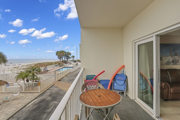 A balcony with a view of the beach, a table, chairs, and a pool.