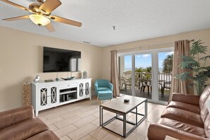 A spacious living room with a ceiling fan, a flat-screen TV, a white cabinet, a blue armchair, a brown leather sofa, and a glass door leading to an outdoor patio with a view of the ocean.