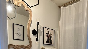 Ein Badezimmer mit weißem Waschbecken, Waschtisch und Toilette. An den Wänden befinden sich Bilder in Rahmen, ein Spiegel mit aufwendigem Rahmen und ein Duschvorhang.