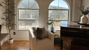 Ein Zimmer mit zwei großen Bogenfenstern, einem Klavier, einer Couch mit Kissen und einer Topfpflanze.