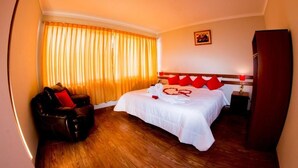 A hotel room with a wooden floor, a bed with white bedding and red pillows, a black leather chair, and a window with orange curtains. - CUSCO HOUSE HOTEL (Cusco)