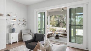 Una sala de estar moderna con un sofá gris, un sillón blanco y una mesa de centro redonda. Hay estantes con objetos decorativos y un armario blanco con una lámpara. La habitación tiene grandes puertas de vidrio que dan a un patio exterior con un comedor y vistas a árboles.