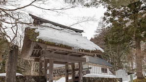Une construction traditionnelle en bois, avec un toit enneigé, entourée d’arbres et d’un mur de pierre.