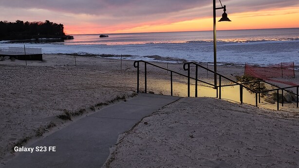 A beach with a walkway leading to the water, a lamp post, and a fence.