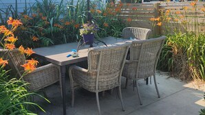 An outdoor patio with a table and wicker chairs, surrounded by plants and flowers.