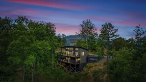 Una casa moderna de dos pisos con un balcón, rodeada de un bosque denso.
