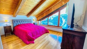 A bedroom with a large bed, wooden ceiling beams, a window with a view, and a TV on a stand.