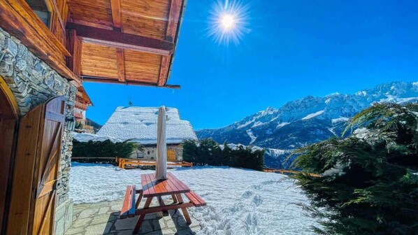 A wooden cabin with a stone exterior, a wooden deck, and a view of snow-covered mountains.