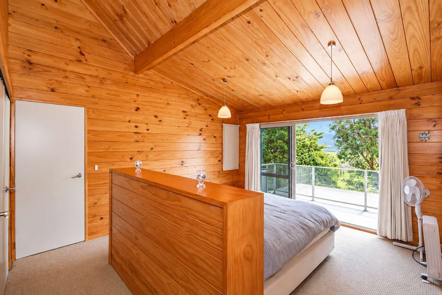 Une chambre dans un chalet en bois, comprenant un lit, une commode, une porte-fenêtre coulissante donnant sur un balcon et un ventilateur de plafond.