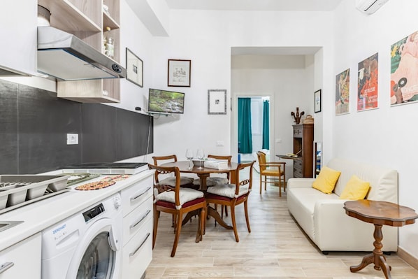 A modern kitchen with a washing machine, dining area with chairs, a sofa, and artwork on the walls.