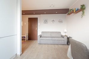 A modern living room with a grey sofa, a small table with a lamp, and a wooden shelf with plants. - GuestHost - Comfortable and modern 45sqm apartment, able to comfortably accommodate up to 4 people, located in the basement of a building WITHOUT a lift (steps to access it). The apartment is located in a strategic position, just a 5-minute walk from the (Milan)