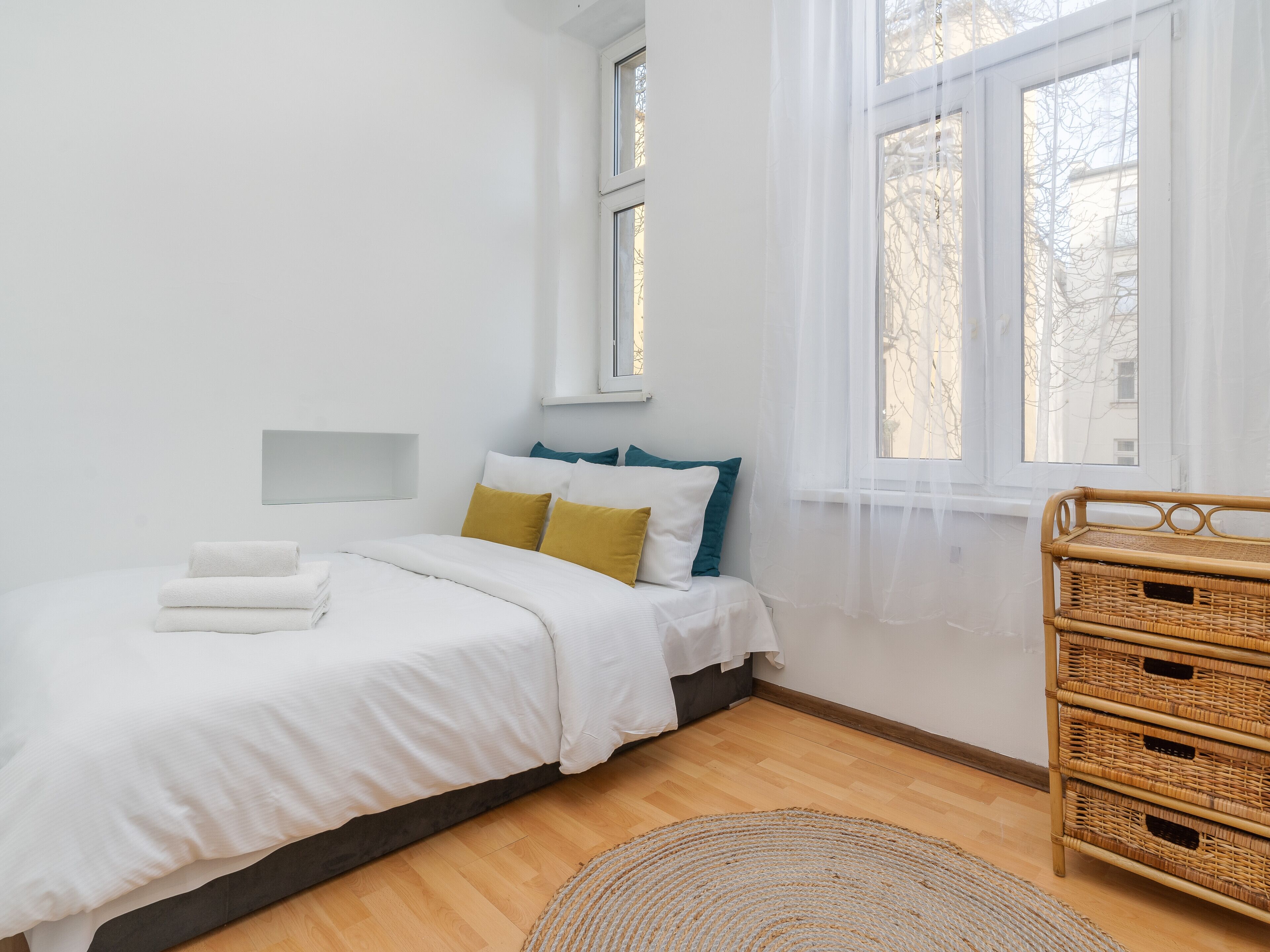 Une chambre à coucher comprenant un lit, une table de chevet, une fenêtre et un meuble de rangement en osier.