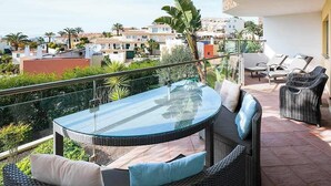 A balcony with a glass table and wicker furniture, overlooking a residential area with palm trees and houses.