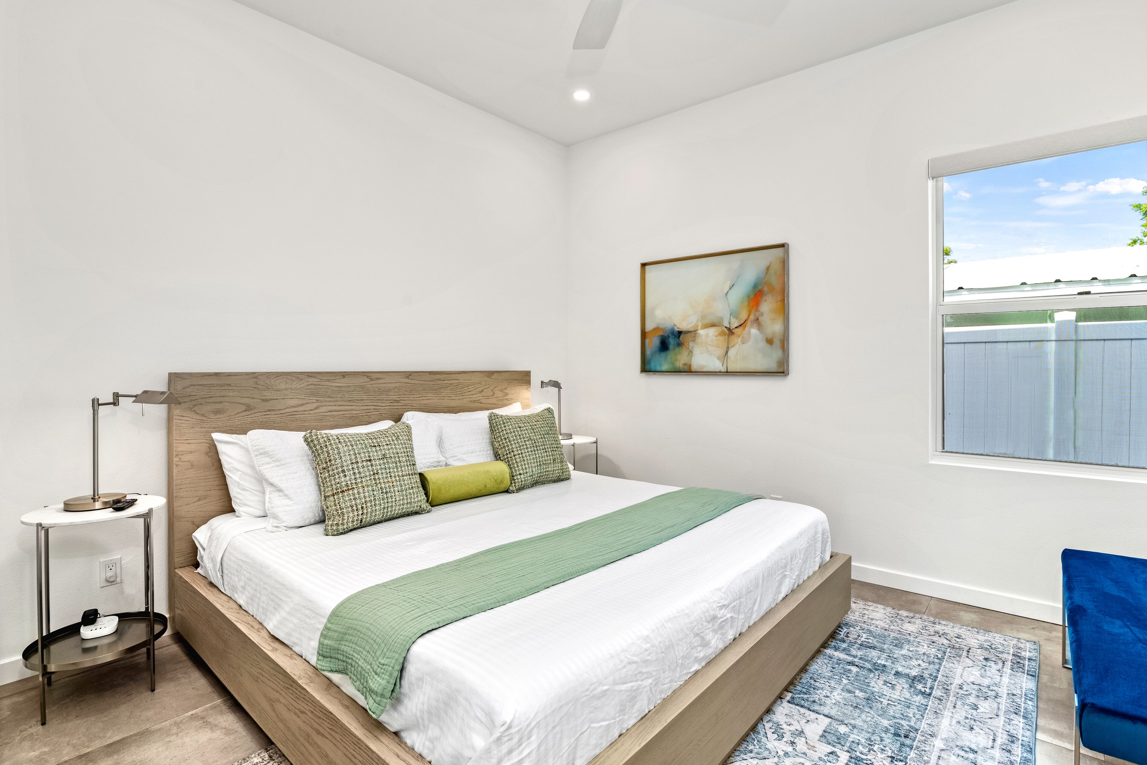 Une chambre à coucher avec un lit en bois, du linge de lit blanc, un plaid vert, un tapis bleu et une fenêtre donnant sur une clôture.