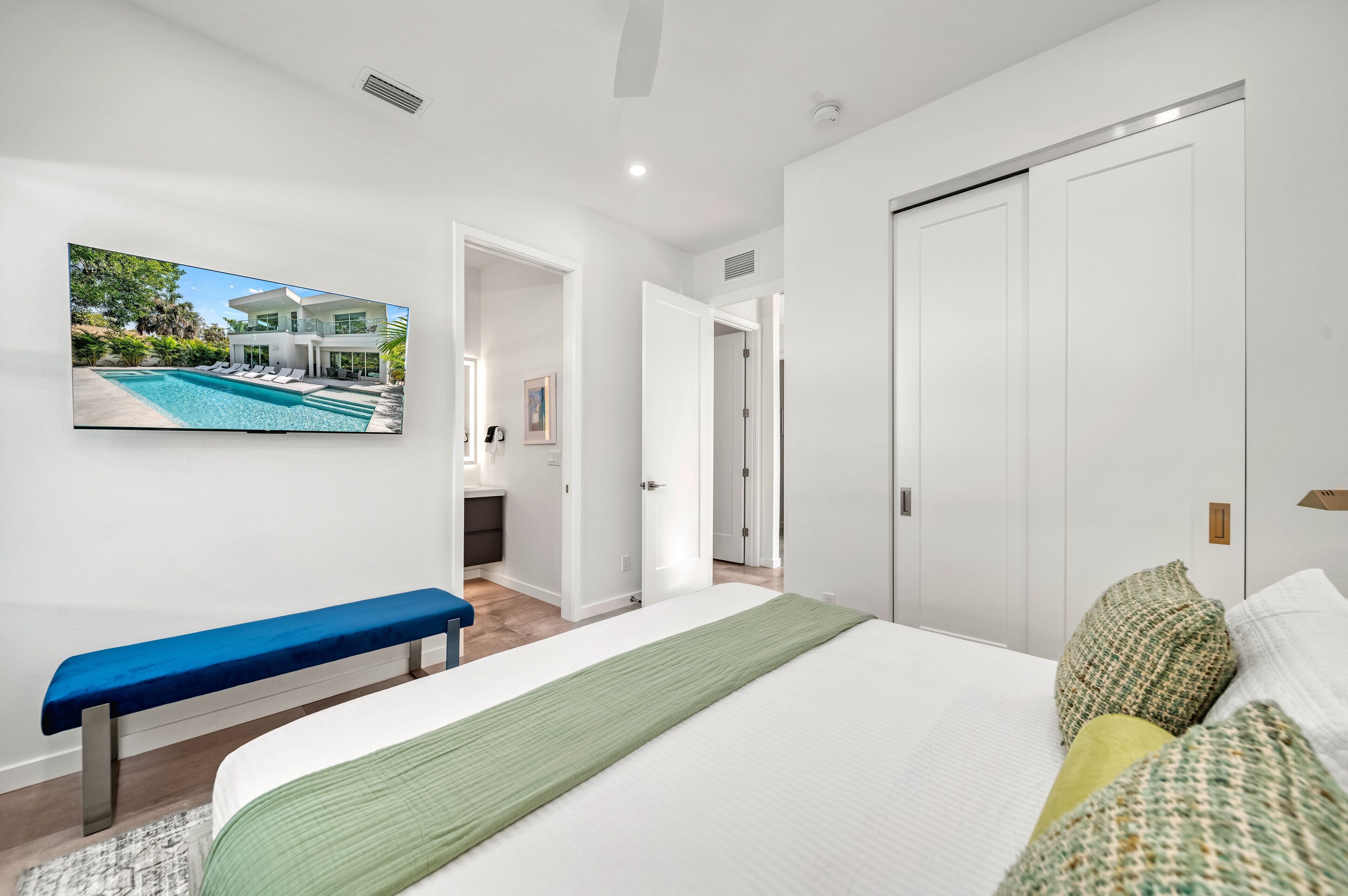Un dormitorio moderno con una cama grande, un banco, un televisor que muestra vista a una piscina y puertas blancas.
