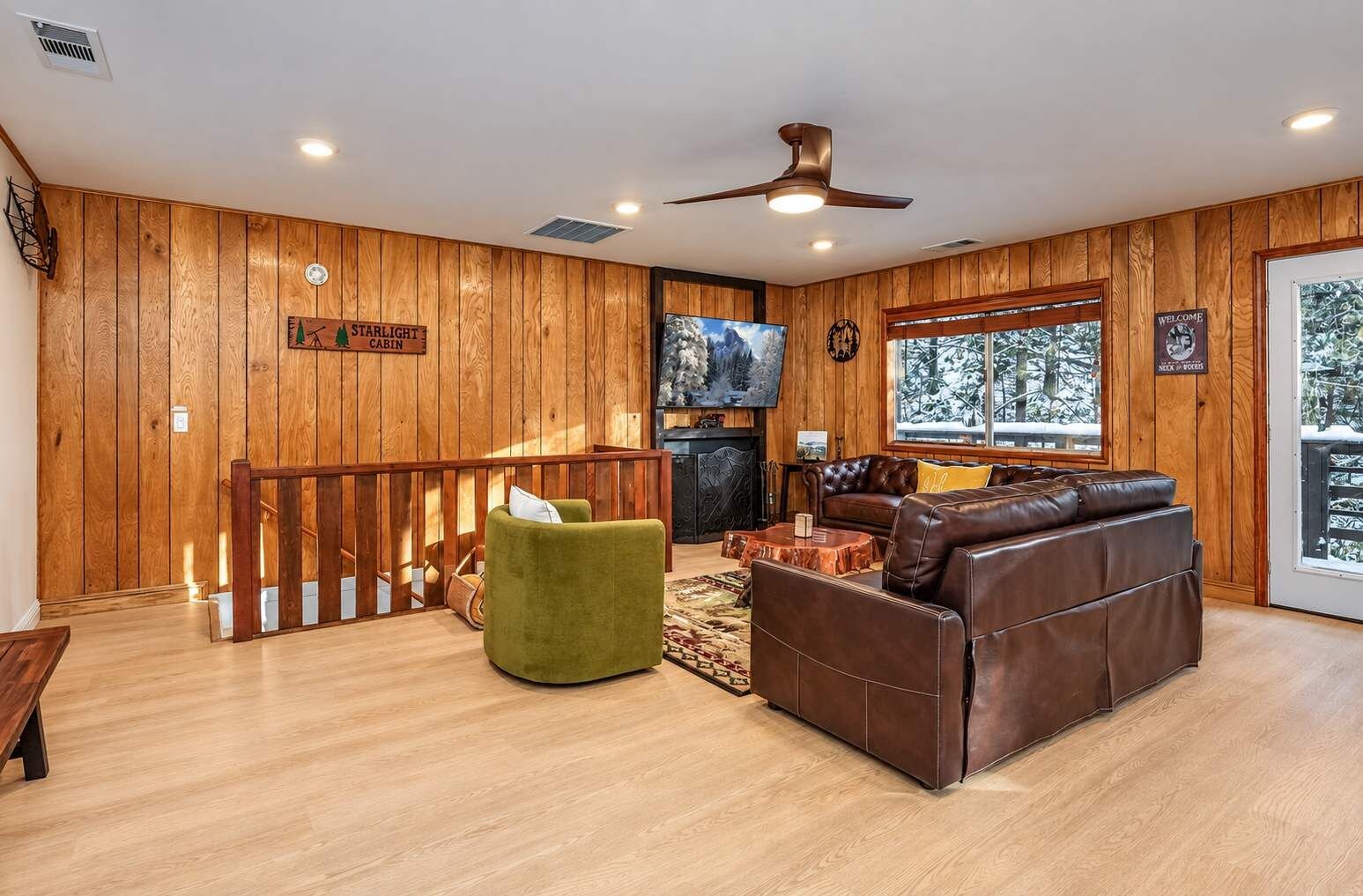 A cabin interior with a wooden floor, a brown leather sofa, a green armchair, a wooden railing, and a fireplace.