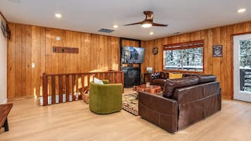 A cabin interior with a wooden floor, a brown leather sofa, a green armchair, a wooden railing, and a fireplace.