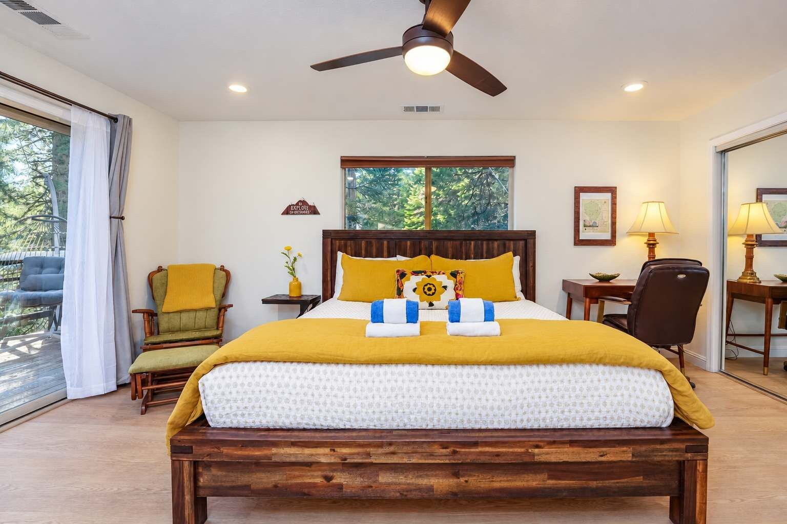 A bedroom with a bed, a desk, a chair, a ceiling fan, and a window with a view of trees.