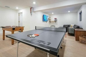 Una mesa de ping pong en una habitación con una mesa de fútbol de mesa, un sofá y un televisor montado en la pared.