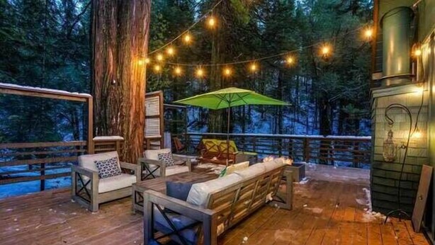 A wooden deck with outdoor seating, string lights, and a large tree in the background.