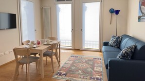 Uma sala de estar moderna com um sofá, uma mesa de jantar com cadeiras, uma TV na parede e grandes janelas com persianas.