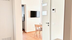 Quarto de hotel moderno com piso de madeira, área de refeições com mesa e cadeiras, TV de tela plana fixada na parede e um guarda-roupa branco com espelho.