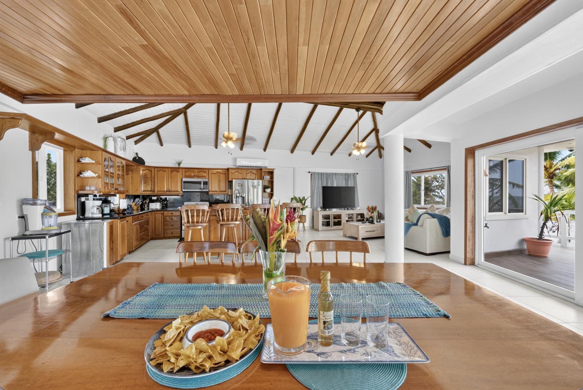 A spacious living area with a wooden ceiling, a dining table with snacks and drinks, and a kitchen in the background.