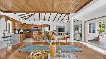A spacious living area with a wooden ceiling, a dining table with snacks and drinks, and a kitchen in the background.