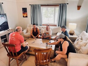 Un groupe de personnes est assis autour d’une table basse en bois dans un salon, jouant aux cartes. On y voit également un téléviseur, une lampe et un tableau accroché au mur.