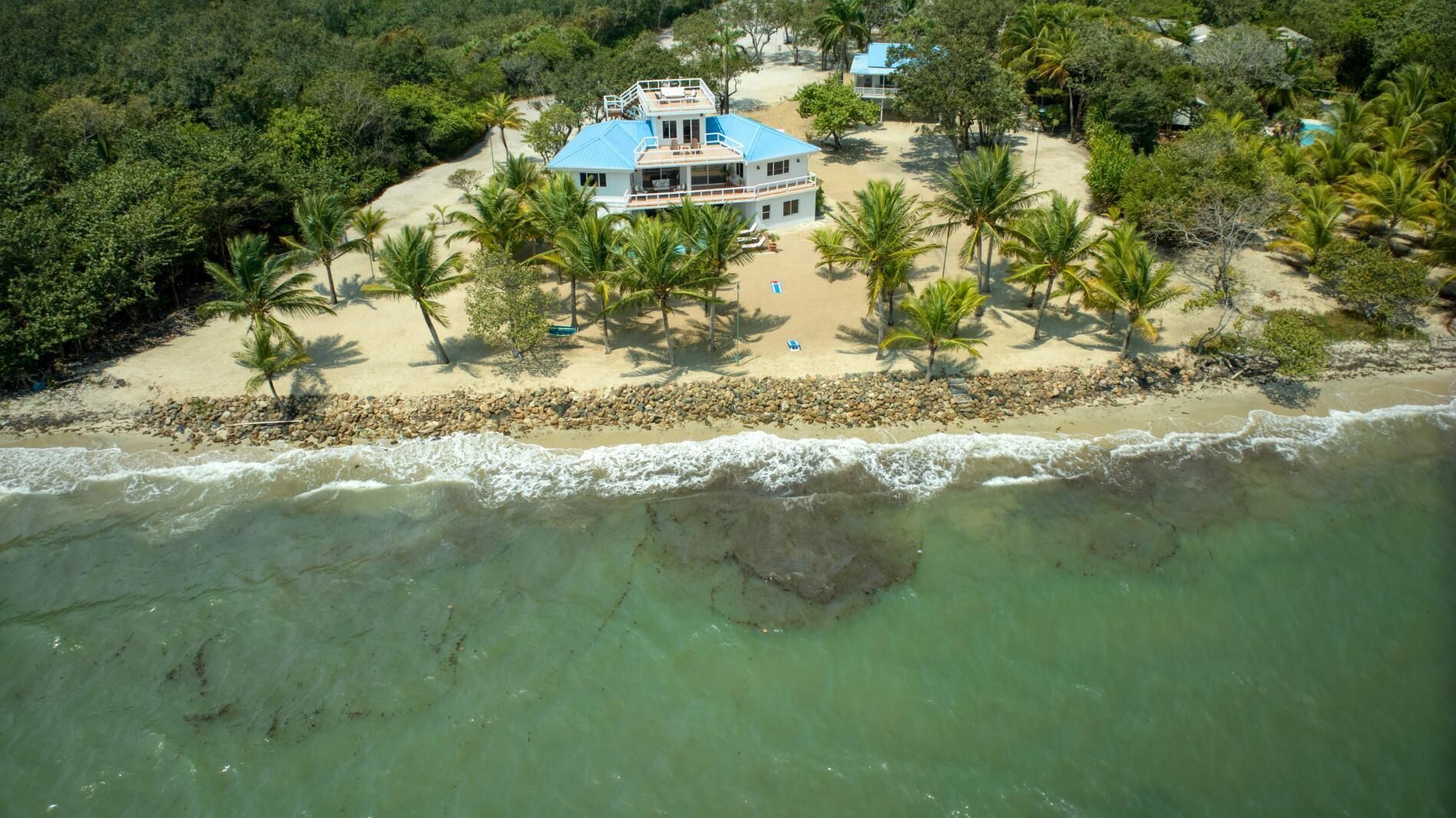 An aerial view of a beachfront property with a large house, multiple palm trees, and a rocky shoreline.