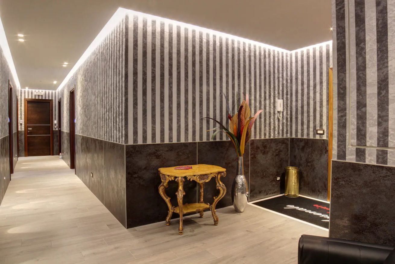 Um lobby moderno de hotel com piso de mármore, balcão de recepção de madeira, um vaso com plantas decorativas e uma parede listrada em preto e branco.