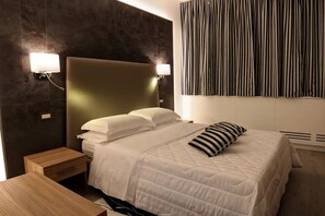 A hotel room with a bed, bedside tables, a headboard, wall-mounted lights, and a striped curtain.