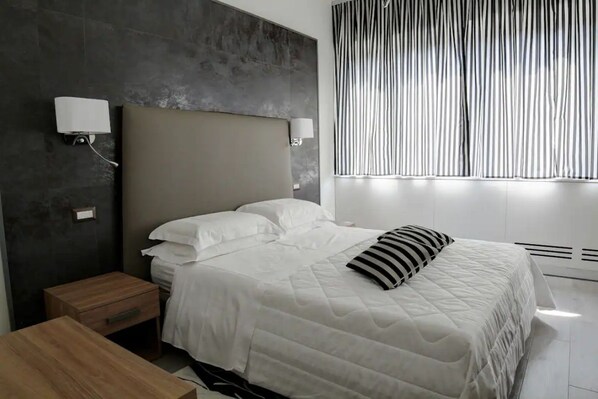 A modern bedroom with a large bed, bedside tables, a nightstand, a wall-mounted lamp, and a window with vertical blinds.