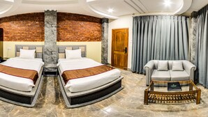 Quarto de hotel com parede de destaque em tijolo, cama com duas almofadas, poltrona cinza, mesa pequena e televisão na parede.
