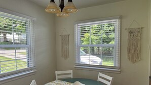 A dining area with a round table, white chairs, and a patterned tablecloth. There are two windows with blinds, a macrame wall hanging, and a chandelier.