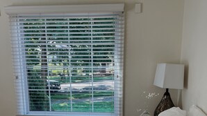 A bedroom with a bed, a window with blinds, a lamp, and a view of greenery outside.