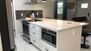 A modern kitchen with white cabinetry, a central island, and a built-in oven. There are pendant lights above the island and a window with curtains. - Cozy ,Tranquil 3-bedroom in Spring Hill with WiFi, AC ; home away from home.  (Spring Hill)