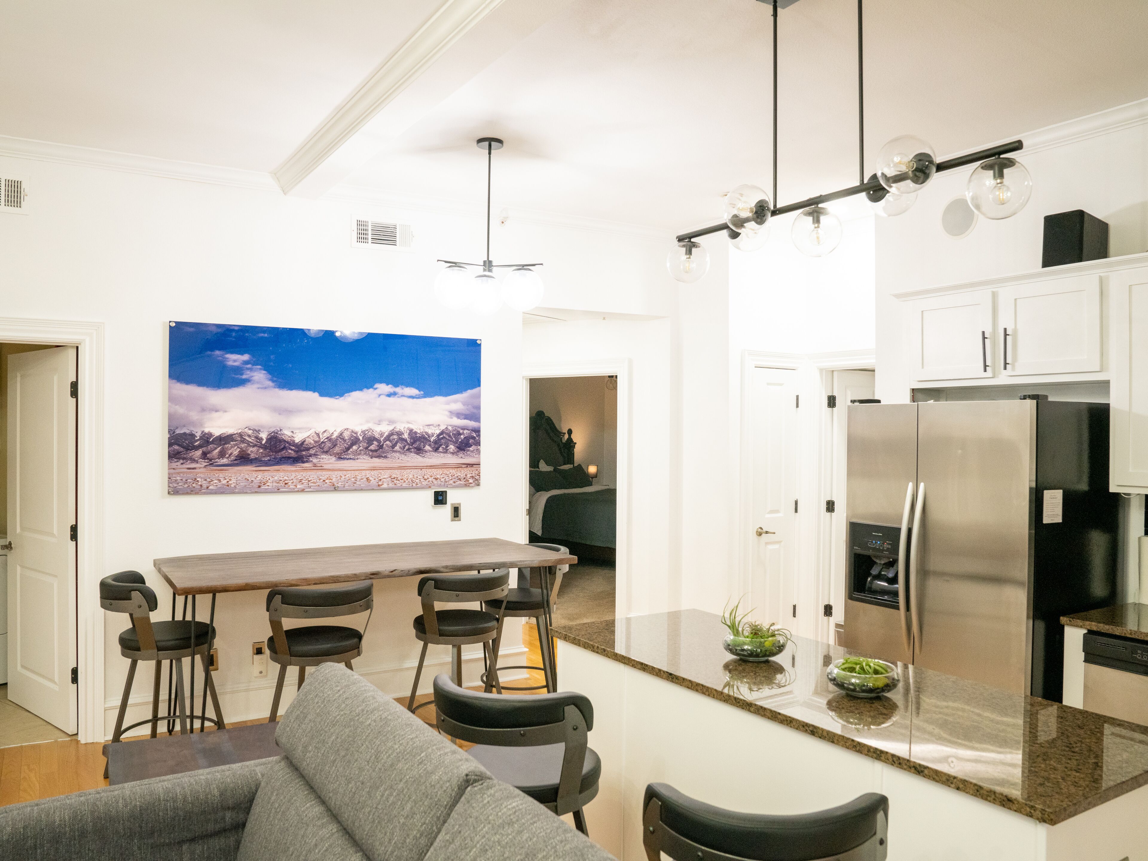 A modern kitchen with a stainless steel refrigerator, a bar area with stools, and a large painting of a mountain landscape on the wall.