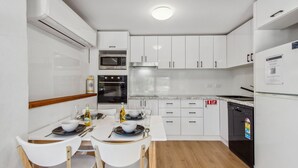 Cocina moderna con armarios blancos, horno empotrado, microondas y lavavajillas. Hay una mesa de comedor con sillas y una pequeña isla con un bol y una botella.